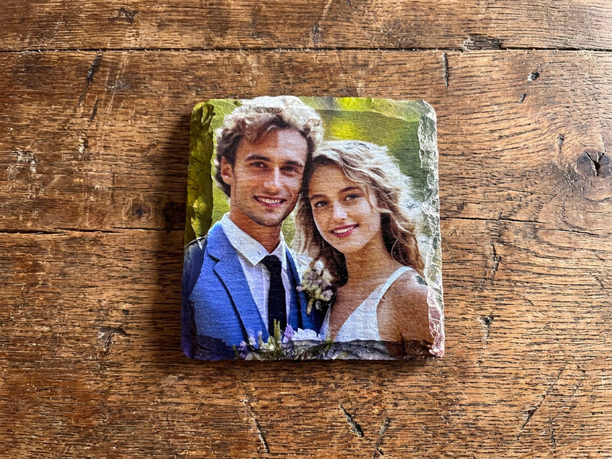 A square stone coaster featuring a custom printed photo of a smiling couple, displayed on a rustic wooden tabletop.