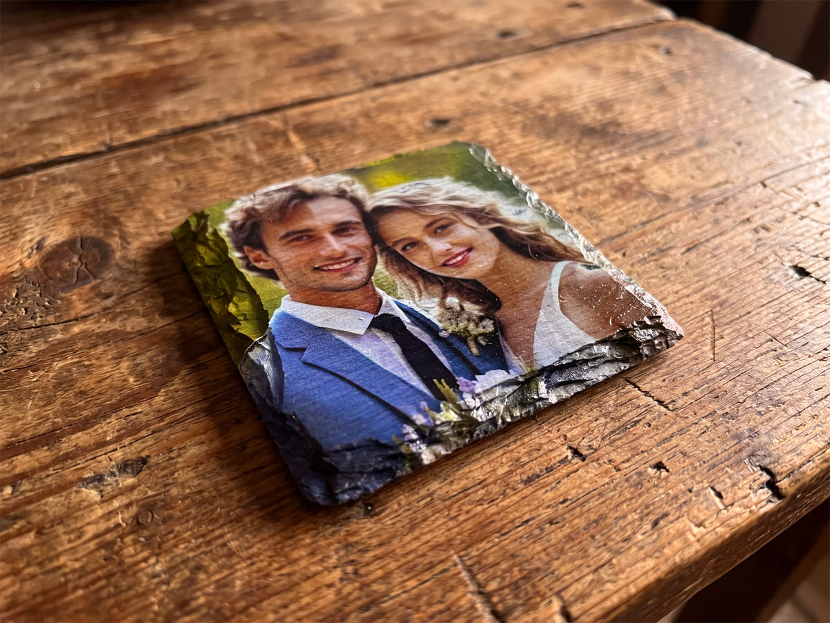 A square custom photo slate coaster featuring a printed wedding photograph of a couple, resting on a rustic wooden table.